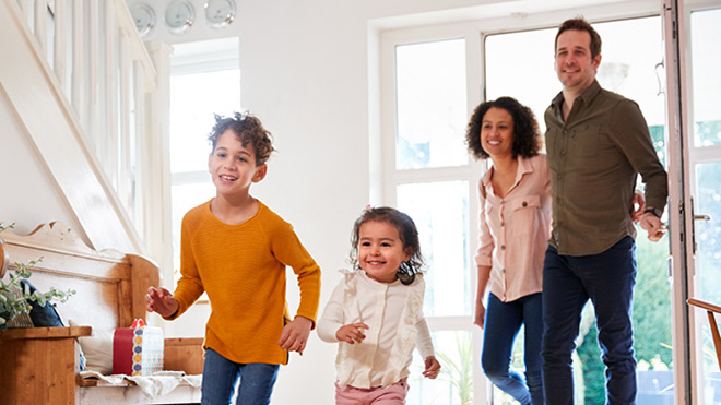 A family of four enters a home with excitement. The parents walk arm in arm, with the father holding the open door. Their two smiling children lead the way into the entryway.