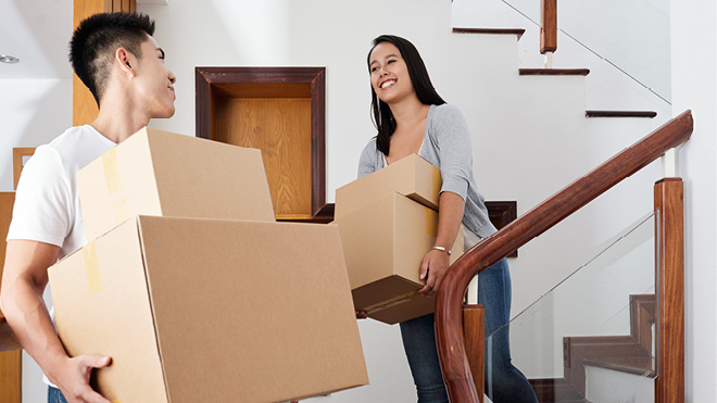 Two young people smile at each other as they walk down the stairs inside a home or apartment while holding a pair of moving boxes in each of their hands. 