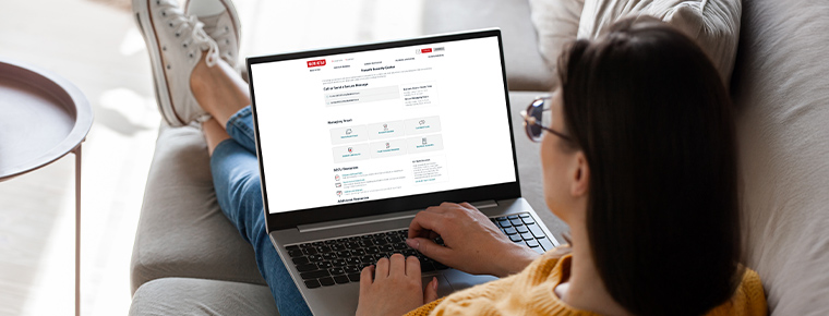 A person sits cross-legged on a couch with their back to the camera. A laptop rests on their lap, displaying a Fraud & Security webpage.