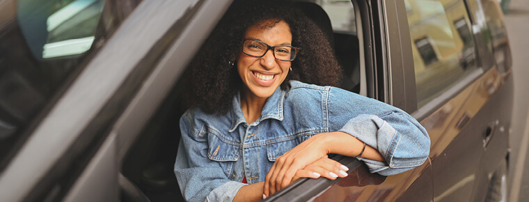 A woman with curly hair and glasses smiling while leaning out of the driver’s side window of a car, wearing a denim jacket.