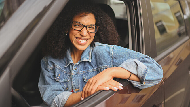 A woman with curly hair and glasses smiling while leaning out of the driver’s side window of a car, wearing a denim jacket.