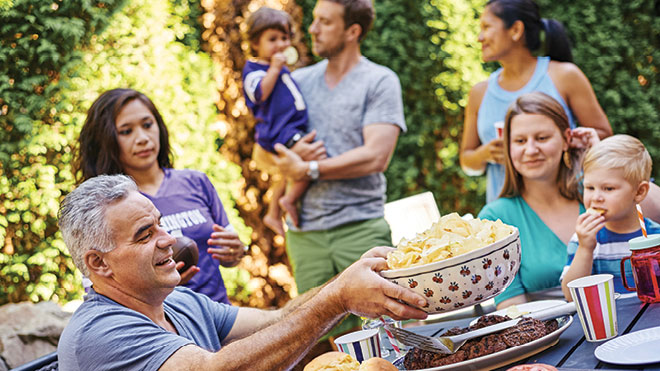 A family enjoys a meal together at a picnic table. A man is seated passing out chips and other family members are gathered and seated around the table with burgers.