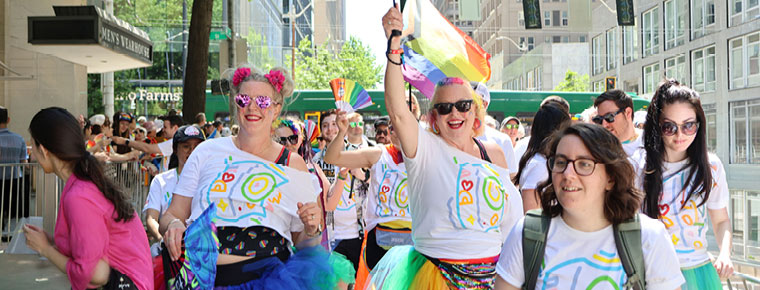 BECU employees march in the Seattle Pride Parade wearing matching white tee-shirts with a colorful design. Two people at the center of the photo are wearing rainbow colored tutus, have colorful hair and are carrying a rainbow umbrella. A Pride flag waves in the background.