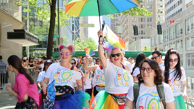 BECU employees march in the Seattle Pride Parade wearing matching white tee-shirts with a colorful design. Two people at the center of the photo are wearing rainbow colored tutus, have colorful hair and are carrying a rainbow umbrella. A Pride flag waves in the background.