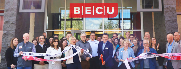 About 30 people stand outside in front of tall glass doors. A man in the middle of the photo holding oversized scissors has cut through large red and white ribbons. A red BECU logo is centered at the top of the image. 