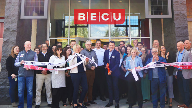 About 30 people stand outside in front of tall glass doors. A man in the middle of the photo holding oversized scissors has cut through large red and white ribbons. A red BECU logo is centered at the top of the image.