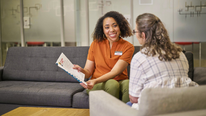 A BECU member consultant shows a brochure to a member. The member consultant is sitting on a couch and smiling at the member who is seated across from her in a cushioned chair facing away from the camera.