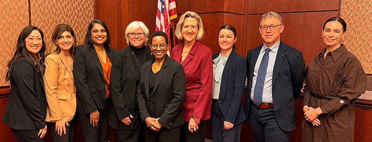 Nine professionals stand together in business attire inside a wood‑paneled government building, with a red carpet and a U.S. flag in the background.