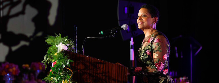 BECU President and CEO Beverly Anderson speaks at a podium during a formal event, with stage lighting and floral decor in the background.