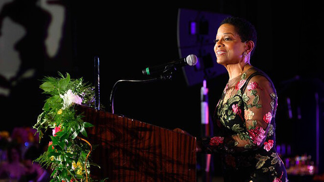 BECU President and CEO Beverly Anderson speaks at a podium during a formal event, with stage lighting and floral decor in the background.