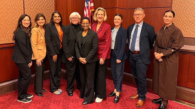 Nine professionals stand together in business attire inside a wood‑paneled government building, with a red carpet and a U.S. flag in the background.