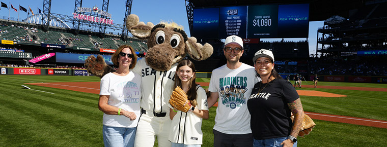 Young baseball fan holding a glove poses with family members and Mariner Moose mascot on the field at T-Mobile stadium.