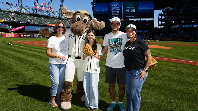 Young baseball fan holding a glove poses with family members and Mariner Moose mascot on the field at T-Mobile stadium.