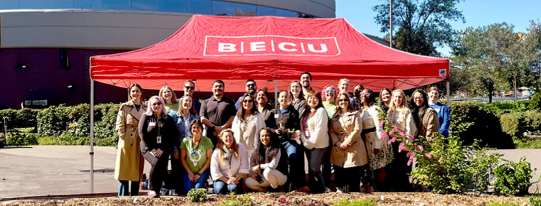 More than 25 BECU employees stand and kneel in the sun in front of a red tent with the white BECU logo that is set up on a sidewalk. Bushes, trees and the Tukwila Financial Center are in the background.
