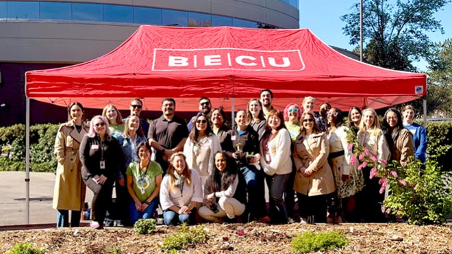More than 25 BECU employees stand and kneel in the sun in front of a red tent with the white BECU logo that is set up on a sidewalk. Bushes, trees and the Tukwila Financial Center are in the background.