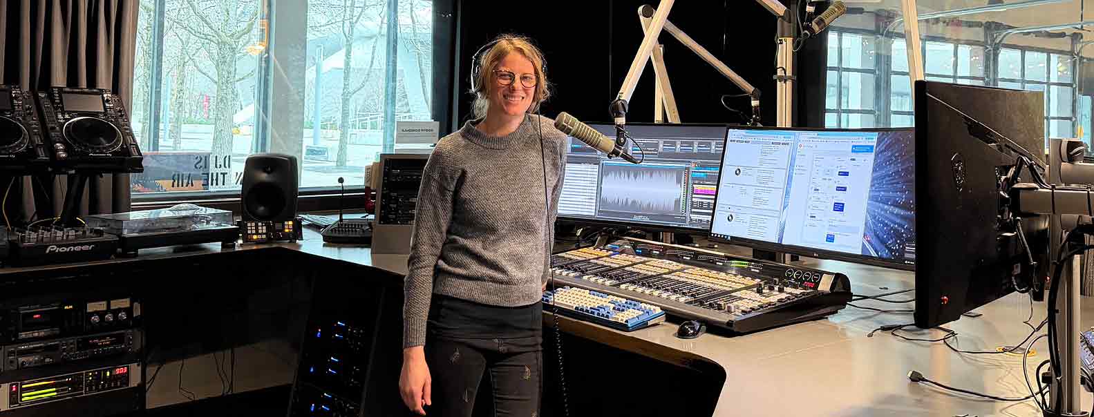 A woman stands in a professional radio studio beside a mixing console, wearing headphones and positioned near a broadcast microphone, with multiple computer monitors displaying audio and messaging software.