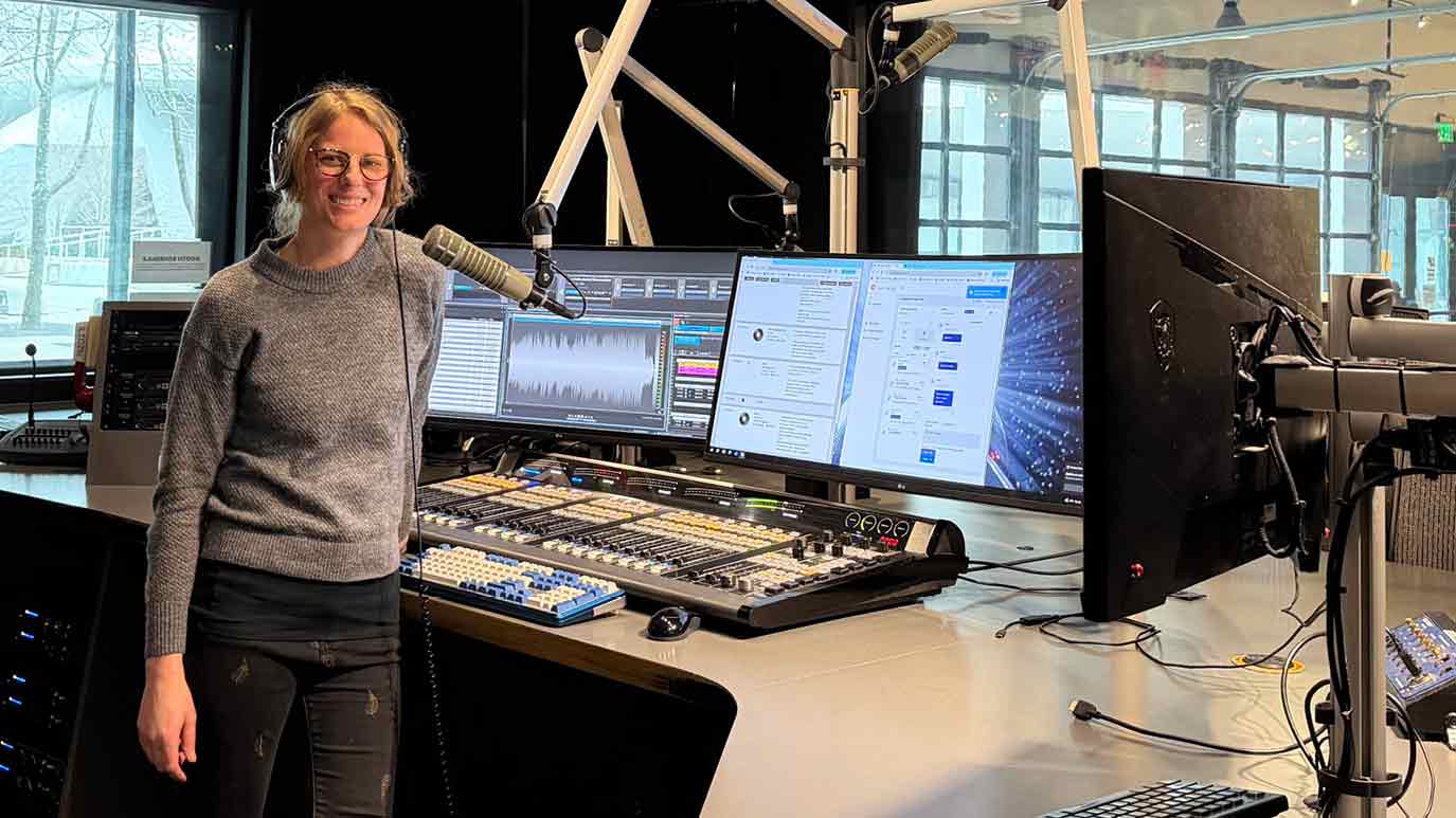 A woman stands in a professional radio studio beside a mixing console, wearing headphones and positioned near a broadcast microphone, with multiple computer monitors displaying audio and messaging software.
