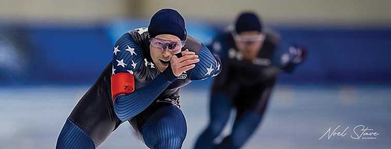 Cooper McLeod speedskating in a blue racing suit, leading a race on an indoor rink.