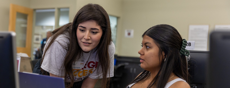 A person wearing a College Success Foundation T-shirt leans toward a seated person as they look at a computer screen together.