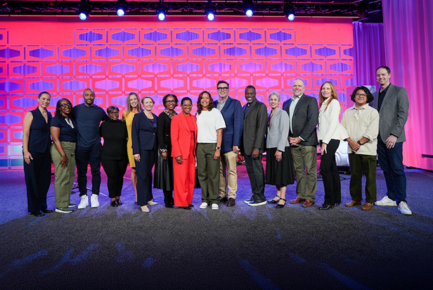Sixteen people representing BECU, BECU Foundation, BECU Board of Directors and nonprofit partners stand together on a stage with a pink and purple backdrop.