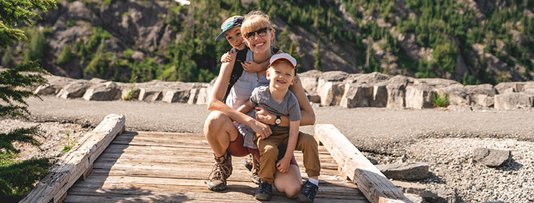 The image shows a smiling woman with two young children on a wooden footbridge during a hike. They're dressed for outdoor travel, with rocky terrain, trees and mountains behind them.
