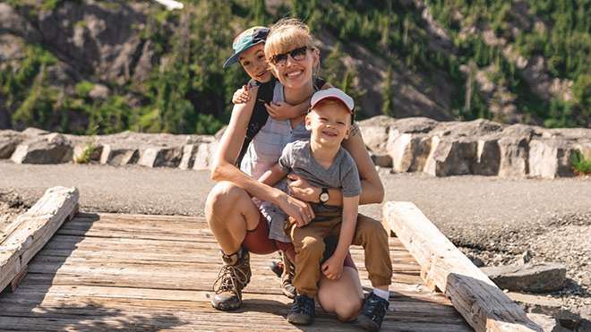 The image shows a smiling woman with two young children on a wooden footbridge during a hike. They're dressed for outdoor travel, with rocky terrain, trees and mountains behind them.
