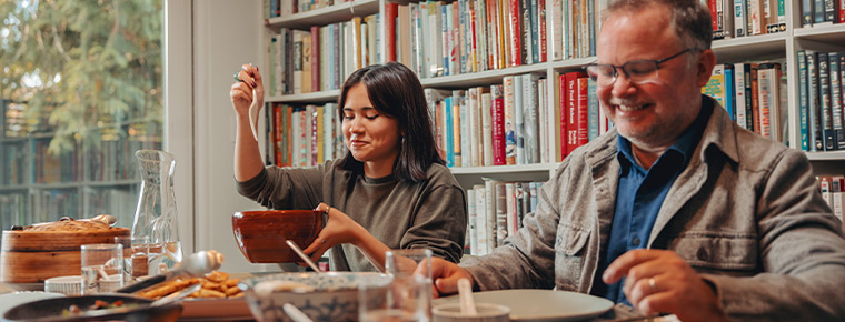 Two people sit at a dining room table, sharing food, with bookshelves behind them.