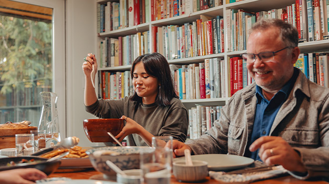 Two people sit at a dining room table, sharing food, with bookshelves behind them.