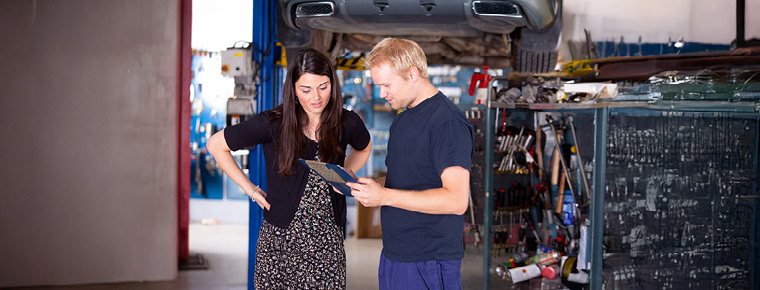 Two people stand in an auto repair shop reviewing a clipboard beneath a car on a lift.