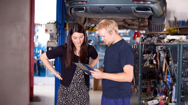 Two people stand in an auto repair shop reviewing a clipboard beneath a car on a lift.