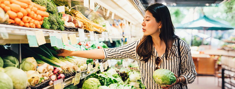 Person reaching for vegetables in a grocery store produce aisle, holding a head of lettuce with shelves of fresh greens and carrots nearby.