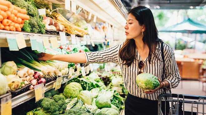 Person reaching for vegetables in a grocery store produce aisle, holding a head of lettuce with shelves of fresh greens and carrots nearby.