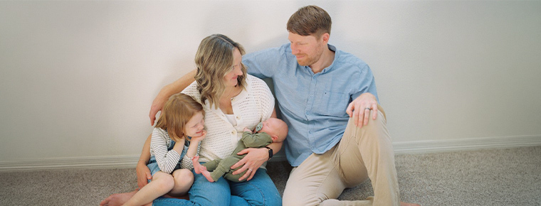 Two adults sit on the floor with a young child while one adult cradles a newborn, all seated closely together.