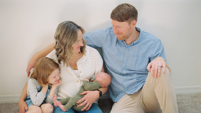 Two adults sit on the floor with a young child while one adult cradles a newborn, all seated closely together.