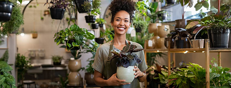 A woman wearing an apron holding a potted plant inside an indoor plant shop, filled with hanging and shelved greenery.