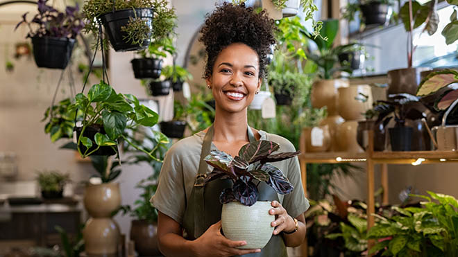 A woman wearing an apron holding a potted plant inside an indoor plant shop, filled with hanging and shelved greenery.