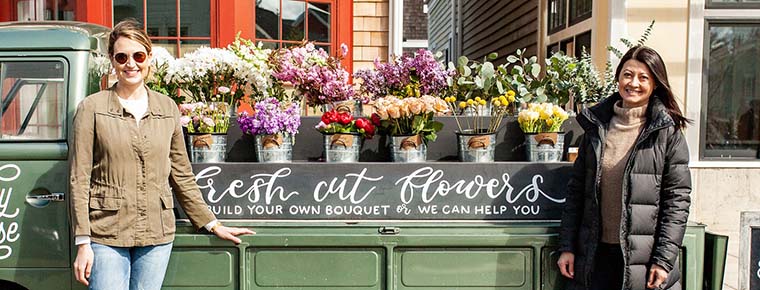 Two small business owners stand outside a flower truck filled with fresh bouquets in front of a local storefront.