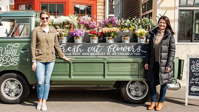 Two small business owners stand outside a flower truck filled with fresh bouquets in front of a local storefront.