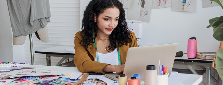 A small business owner works at a desk with a laptop, fabric samples and sewing tools in a creative workspace.