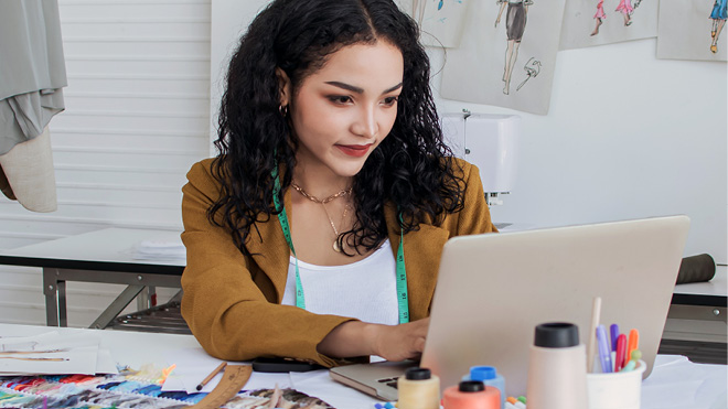 A small business owner works at a desk with a laptop, fabric samples and sewing tools in a creative workspace.