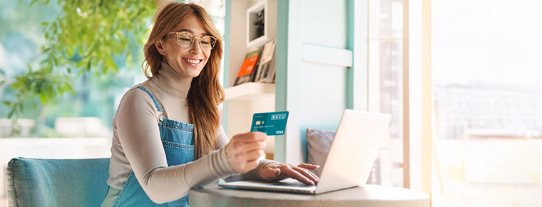 Smiling woman holding a credit card while using a laptop, seated at a table in a sunlit room.