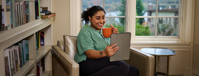 Smiling person seated in an armchair, holding a tablet in one hand and a mug in the other.