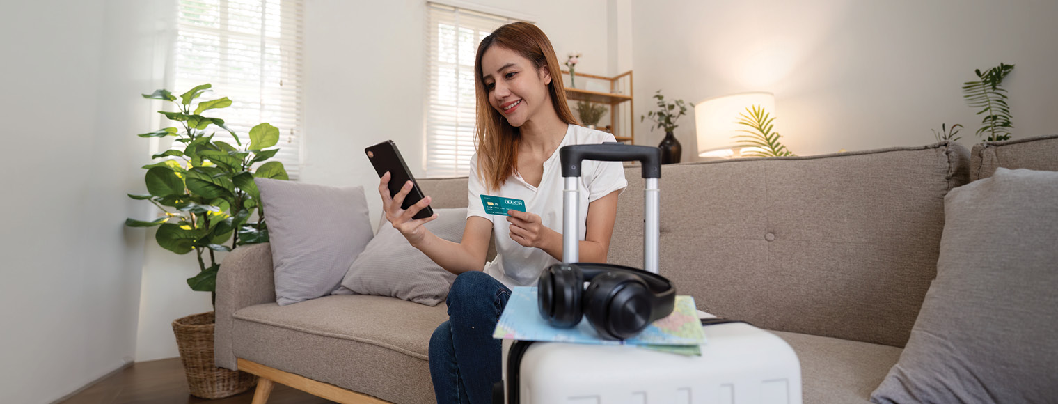 Woman sitting on a sofa, holding a smartphone and credit card beside a suitcase and headphones.