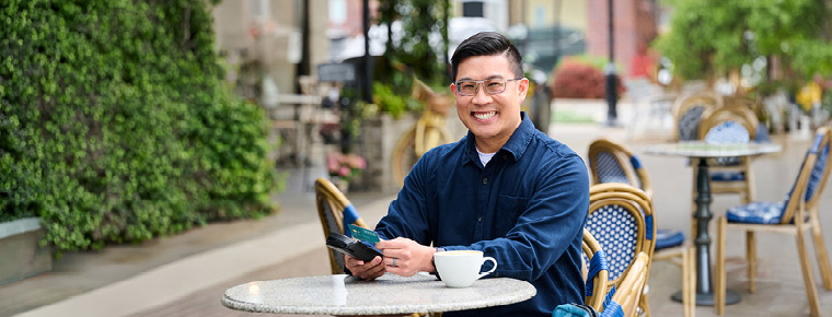 Person seated at an outdoor café table, holding a smartphone and credit card beside a cup of coffee, with café chairs and greenery in the background.