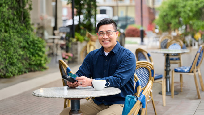 Person seated at an outdoor café table, holding a smartphone and credit card beside a cup of coffee, with café chairs and greenery in the background.