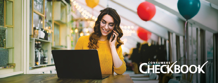 A woman wearing a bright yellow sweater smiles while holding a phone up to her ear and looking at a laptop. She is sitting in a room with an angled ceiling and white beams. Red, yellow and teal balloons and white string lights are in the background. The Consumers' Checkbook logo appears as white text in the lower right corner of the image.