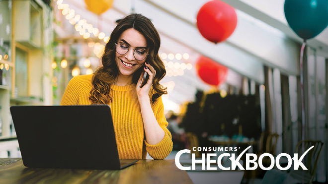 A woman wearing a bright yellow sweater smiles while holding a phone up to her ear and looking at a laptop. She is sitting in a room with an angled ceiling and white beams. Red, yellow and teal balloons and white string lights are in the background. The Consumers' Checkbook logo appears as white text in the lower right corner of the image.