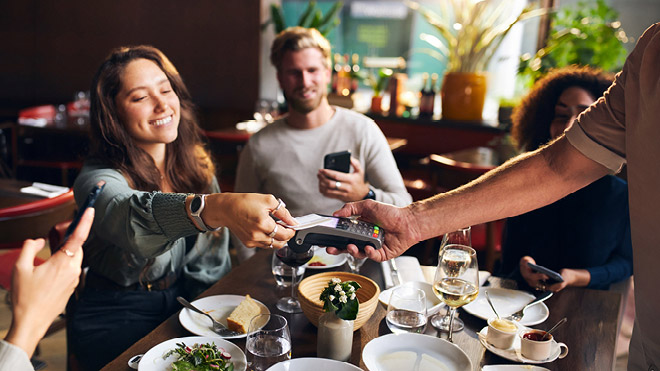 A group of friends sit around a table at a restaurant. A woman smiles as she reaches to pay with her credit card on the card reader held by the server.