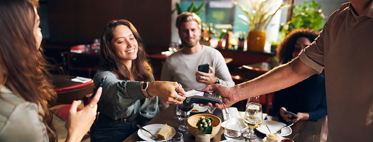 A group of friends sit around a table at a restaurant. A woman smiles as she reaches to pay with her credit card on the card reader held by the server.
