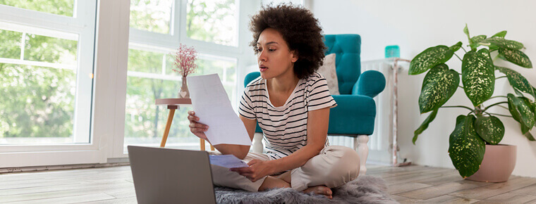 A woman sits cross-legged on a fuzzy rug on a wood floor with her laptop open in front of her. She's looking at a piece of paper she's holding up. A bright turquoise arm chair, houseplant and window are behind her. Trees and sunlight fill the window.